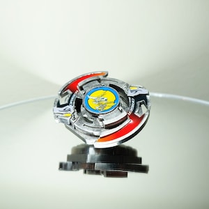 May include: A silver and red Beyblade with a yellow and blue centre. The Beyblade has the word "DRIGER" printed on it.