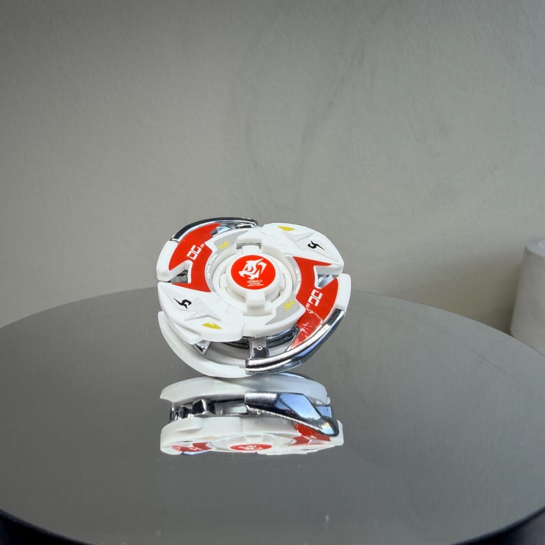 May include: A white and red spinning top toy with a silver metallic rim. The toy has a central design with a red emblem and is reflected on a shiny surface. The top has a circular shape with red and white accents.