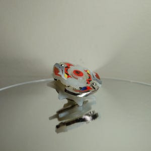 May include: A white, red, yellow, and blue spinning top toy with a silver metal rim. The toy has a circular design with a central emblem. The toy is sitting on a reflective surface.