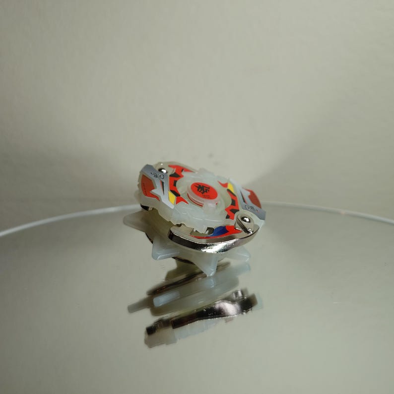 May include: A white, red, yellow, and blue spinning top toy with a silver metal rim. The toy has a circular design with a central emblem. The toy is sitting on a reflective surface.