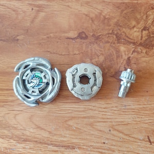 May include: A three-part silver-coloured spinning top toy set. The set comprises the main top with a detailed design, a central piece, and a smaller component. The top features a blue and green emblem. The pieces are arranged on a wooden surface.