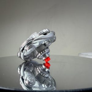 Puede incluir: Una peonza plateada con una punta roja. La peonza tiene un dise&ntilde;o complejo con m&uacute;ltiples capas y detalles. El juguete est&aacute; dise&ntilde;ado para girar y probablemente est&aacute; hecho de pl&aacute;stico o metal.