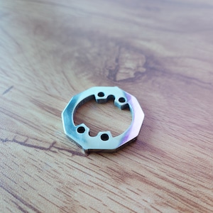 以下が含まれることがあります： 中央に5つの穴が開いた銀色の金属製のリング。リングは木製の表面の上に置かれています。