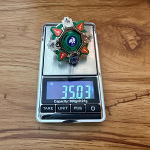 May include: A green, red, and silver toy top rests on a digital scale, displaying a weight of 35.03 grams. The top features a circular design with a central emblem and angular components. The scale has a capacity of 300gx0.01g.