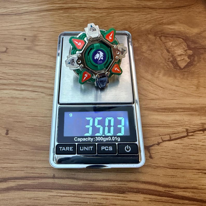 May include: A green, red, and silver toy top rests on a digital scale, displaying a weight of 35.03 grams. The top features a circular design with a central emblem and angular components. The scale has a capacity of 300gx0.01g.