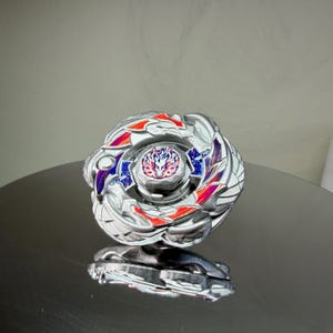 Puede incluir: Un trompo plateado y multicolor con un dise&ntilde;o detallado. La parte superior presenta detalles en rojo, azul y morado, y un emblema central. El juguete est&aacute; dise&ntilde;ado para girar y luchar.