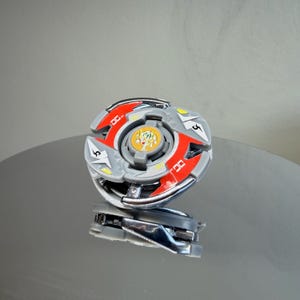 May include: A silver and red toy with a circular design. The toy has a central yellow and green emblem and is reflected in the surface below. The toy has a silver base and red accents.