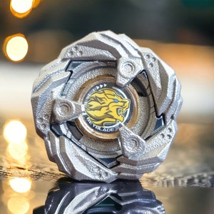 May include: A silver and gold Beyblade toy featuring a lion emblem. The toy has a textured, multi-layered design with the word "Beyblade" printed on the centre. The lion emblem is yellow with blue eyes.
