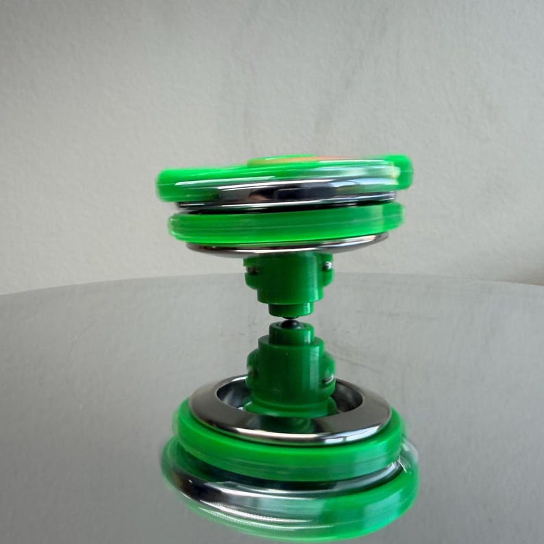 Pu&ograve; includere: Una trottola verde e argento in movimento. Il giocattolo ha un corpo verde con anelli argentati e una base verde. La trottola gira su una superficie riflettente.