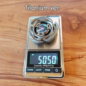 May include: A silver toy top with an intricate design, placed on a digital scale. The scale shows a weight of 50.50 grams. The text "Titanium ver." is above the toy.