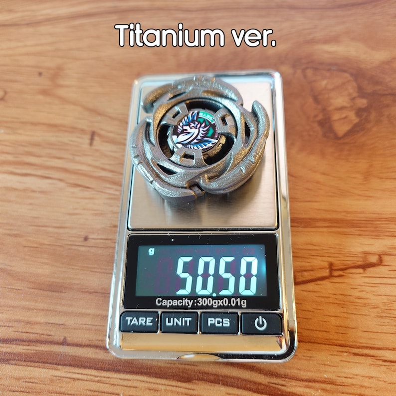May include: A silver toy top with an intricate design, placed on a digital scale. The scale shows a weight of 50.50 grams. The text "Titanium ver." is above the toy.