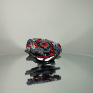 May include: A colorful spinning top toy with a red, gray, and silver design. The top features a central red emblem and is set against a reflective surface, showcasing its intricate details and design.