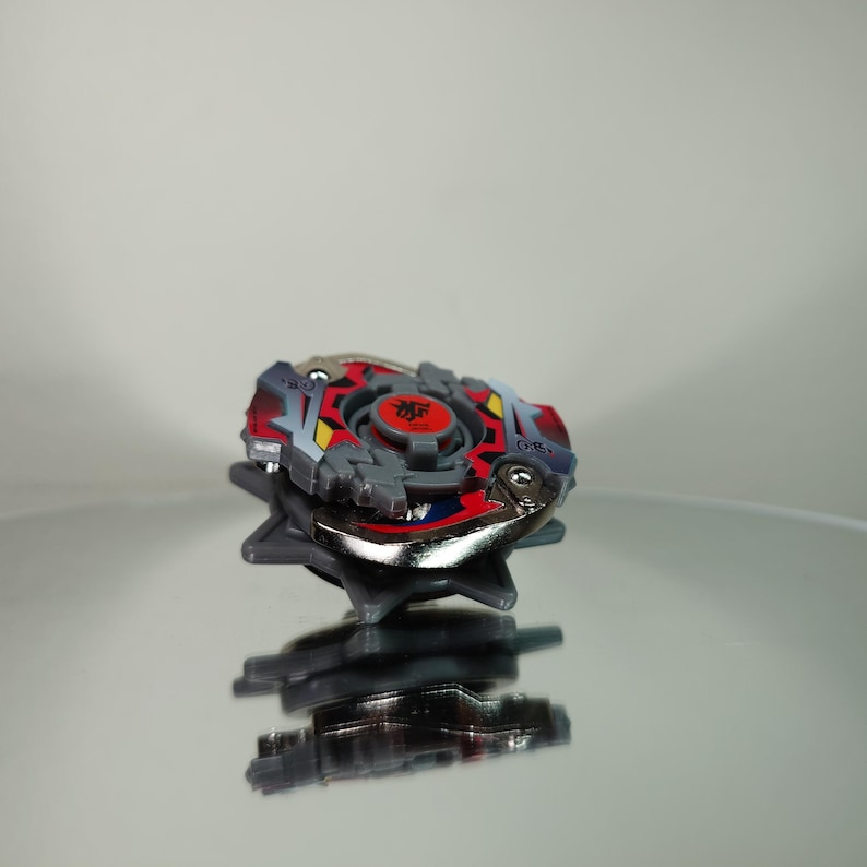 May include: A colorful spinning top toy with a red, gray, and silver design. The top features a central red emblem and is set against a reflective surface, showcasing its intricate details and design.