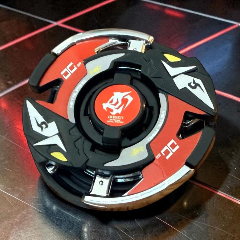 May include: A black, red, and silver toy with a circular design. The toy has a central red circle with white text and a symbol. The outer edges are red with silver accents and white arrow-shaped designs.