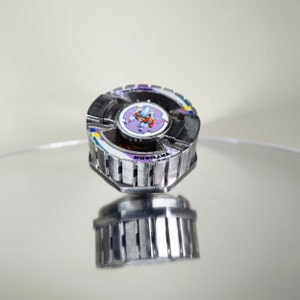 May include: A metallic spinning top toy with a detailed design and multiple colours. The top features a central emblem with a cartoon character and the word "TRYHORN". The toy is designed for spinning and battling.