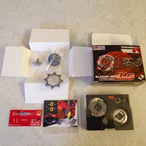 May include: An opened box set of a "Bloody Devil" toy, featuring a red, black, and silver design. The set includes various components, such as a spinning top, a star-shaped piece, and stickers. The box is labeled "HMS G5".