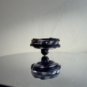 May include: A chrome and black metal spinning top toy. The top has a layered design with a rounded base and a central stem. The top is sitting on a reflective surface, and the background is a solid, neutral color.
