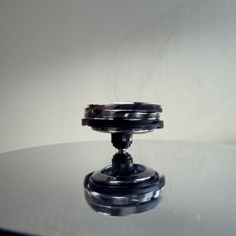 May include: A chrome and black metal spinning top toy. The top has a layered design with a rounded base and a central stem. The top is sitting on a reflective surface, and the background is a solid, neutral color.