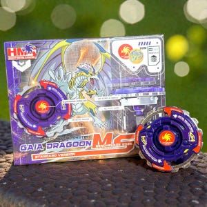 以下が含まれることがあります： 紫と赤のガイアドラグーンベンチブレード2025おもちゃセット。箱には、黄色と青のアクセントが付いたドラゴンのイラストが描かれています。おもちゃはヘビーメタルシステムの標準バージョンです。