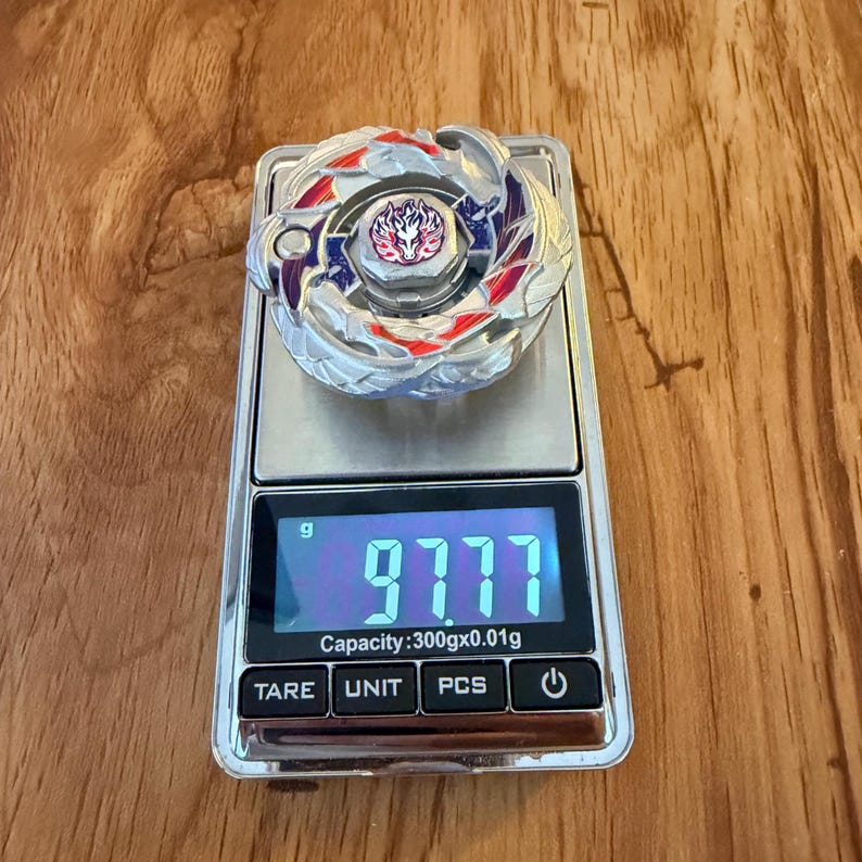 Puede incluir: Un giroscopio de juguete plateado y rojo est&aacute; colocado sobre una balanza digital, mostrando un peso de 97,77 gramos. El giroscopio tiene un dise&ntilde;o central con detalles rojos y morados. La balanza tiene botones para TARE, UNIT, PCS y encendido/apagado.