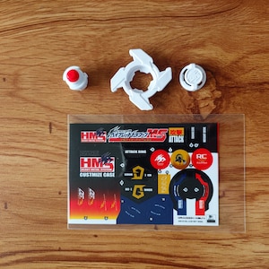 May include: A white plastic Beyblade with a red and black sticker sheet. The sticker sheet has the text "HM HEAVY METAL SYSTEM" and "ATTACK RING".