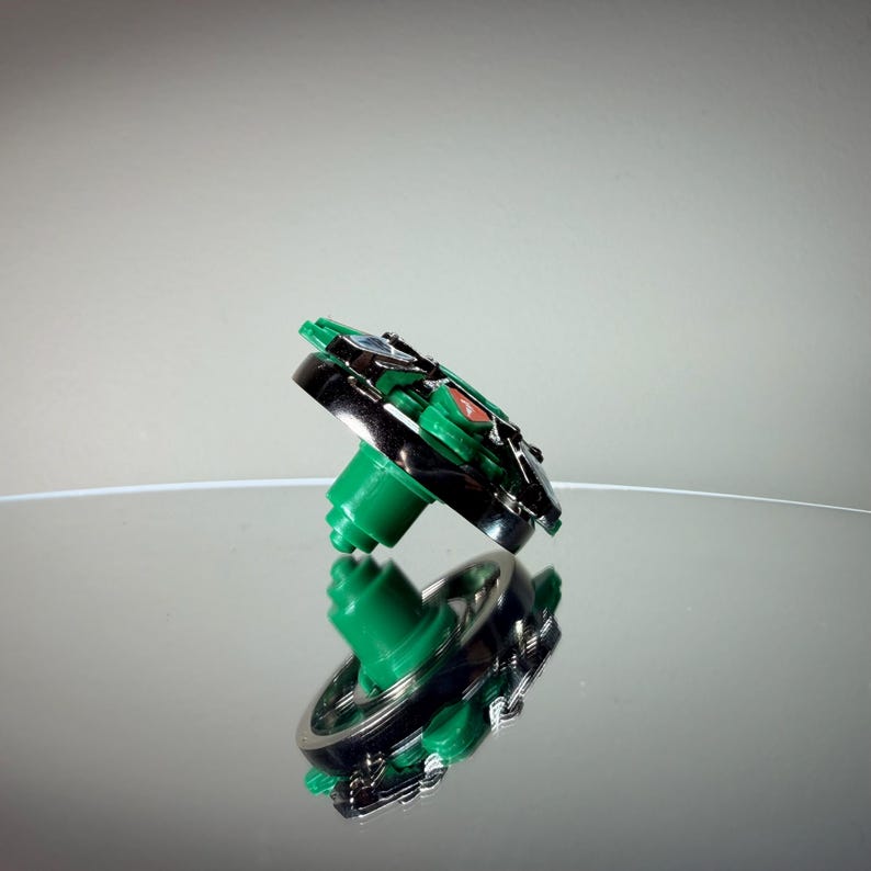 May include: A green and silver spinning top toy with a black rim and a green base. The toy is reflected on a shiny surface. The top has a circular design with red and blue accents.