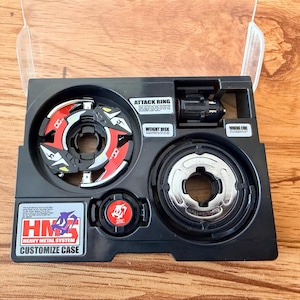May include: A black plastic case containing a red, white, and black spinning top toy. The case is labeled with the text "HMS Heavy Metal System Customize Case". The case also contains the parts "Attack Ring", "Weight Disk", and "Spinning Core".