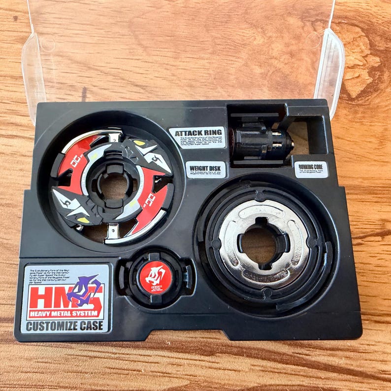 May include: A black plastic case containing a red, white, and black spinning top toy. The case is labeled with the text "HMS Heavy Metal System Customize Case". The case also contains the parts "Attack Ring", "Weight Disk", and "Spinning Core".