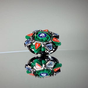 May include: A green, red, silver, and black spinning top toy. The top has a circular design with a central green disc featuring a blue emblem. Red and silver accents are visible around the edges. The toy is reflected on a mirrored surface.