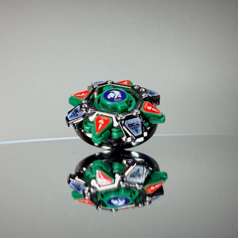 May include: A green, red, silver, and black spinning top toy. The top has a circular design with a central green disc featuring a blue emblem. Red and silver accents are visible around the edges. The toy is reflected on a mirrored surface.
