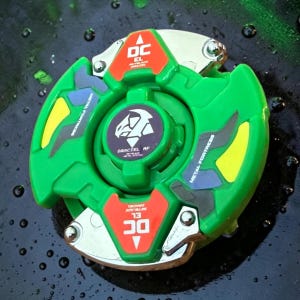 Pu&ograve; includere: Un giocattolo verde e argento con la scritta "DC" e "METAL FORTRESS". Il giocattolo ha un design circolare con un emblema centrale ed &egrave; probabilmente una trottola o un giocattolo simile.