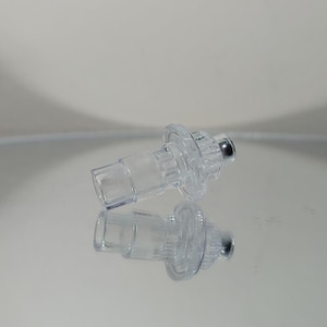 May include: Two clear plastic medical components with a cylindrical shape and a small, silver-colored ball at the end. The components are reflected on a shiny surface, creating a mirrored image. The design includes a circular flange.