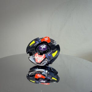 Pu&ograve; includere: Un giocattolo nero e argento con accenti rossi e gialli. Il giocattolo ha un design circolare con le lettere "DC" in rosso. Il centro del giocattolo ha un emblema viola. Il giocattolo si riflette su una superficie lucida.