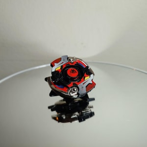 May include: A colorful spinning top toy with a red, black, silver, yellow, and blue design. The toy is on a reflective surface. The top has a circular center with a red emblem and is surrounded by a multi-colored design.