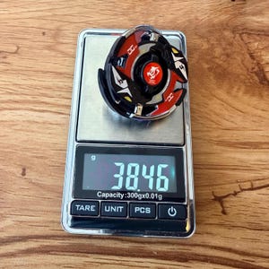 May include: A black, red, and yellow spinning top toy rests on a digital scale. The scale displays a weight of 38.46 grams. The toy has a circular design with a central emblem.