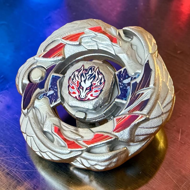 Puede incluir: Un juguete plateado con detalles rojos y azules, con un emblema central de un drag&oacute;n estilizado. El dise&ntilde;o circular tiene patrones intrincados y probablemente sea una peonza o una pieza de juego similar. El juguete incorpora detalles en rojo, azul y morado.