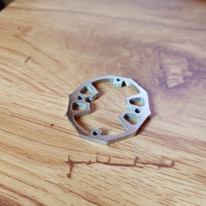 Op de afbeelding: Een zilveren metalen ring met een ronde vorm en meerdere uitsparingen. De ring heeft een gladde, gepolijste afwerking.
