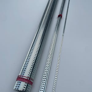 Puede incluir: Un rollo de moldura de metal plateado con agujeros a lo largo de los bordes, y tres piezas individuales de la misma moldura. La moldura se mantiene unida con gomas elásticas rojas.