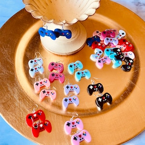 May include: A collection of colorful video game controller earrings. The earrings are made of polymer clay and feature different colors, including blue, pink, red, and black. The earrings are displayed on a gold plate.