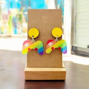May include: A pair of rainbow-colored gamer controller earrings with yellow circle accents. The earrings are displayed on a brown cardboard earring stand.