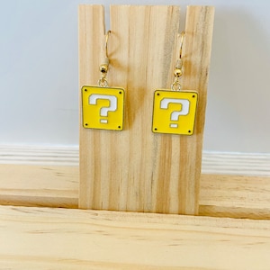 May include: A pair of gold-tone earrings with a yellow square charm featuring a white question mark. The earrings are displayed on a wooden stand.