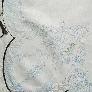 May include: A white linen fabric piece featuring blue ink floral and paisley embroidery patterns. A dark brown stitched border outlines the design, which includes a small rectangular tag reading ALL LINEN MADE IN IRELAND in green text.