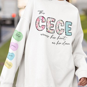 May include: White sweatshirt with the text "This Cece wears her heart on her sleeve" in black. The letters in "Cece" are decorated with floral patterns. The left sleeve has a series of colorful heart-shaped patches with the words "Add", "your", "own", and "text" on them.