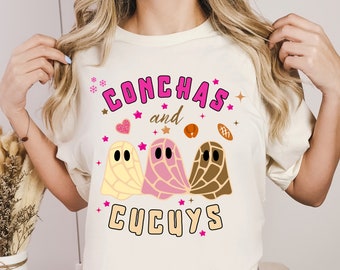 May include: White t-shirt featuring a pink and brown graphic of ghosts and the text "Conchas and Cucuys" in pink lettering.