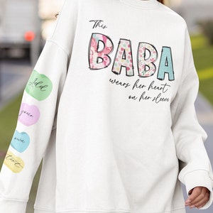 May include: White sweatshirt with the text "This Baba wears her heart on her sleeve" in black script. The word "Baba" is in large, colourful letters with a floral pattern. The sleeve has a series of colourful hearts with the words "Add", "your", "own", and "text" on them.