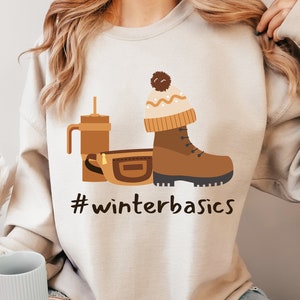 May include: A beige sweatshirt with a graphic design of a brown boot, a brown fanny pack, and a brown to-go cup with a straw. The graphic also includes a brown and white knit hat with a pom-pom. The text "#winterbasics" is printed below the graphic.