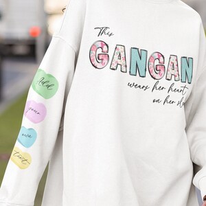 Op de afbeelding: Witte trui met de tekst "This GANGAN wears her heart on her sleeve" en kleurrijke harten op de mouw.