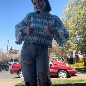 May include: A woman wearing a blue and white crocheted jumper with ruffled sleeves and blue jeans with distressed details. She is giving a thumbs up with both hands.