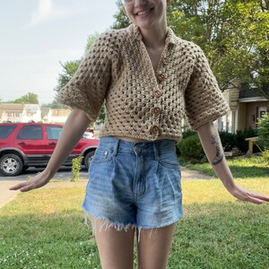 May include: A woman wearing a beige crocheted cropped cardigan with wooden buttons and blue denim shorts.