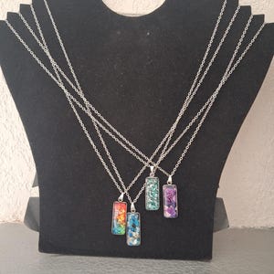 Op de afbeelding: Vier rechthoekige kettingen met hangers en zilverkleurige schakels. Elke hanger heeft een unieke mozaïek-inleg in levendige kleuren zoals rood, oranje, geel, blauw, turquoise en paars, gevat in een gepolijst zilverkleurig metalen frame.